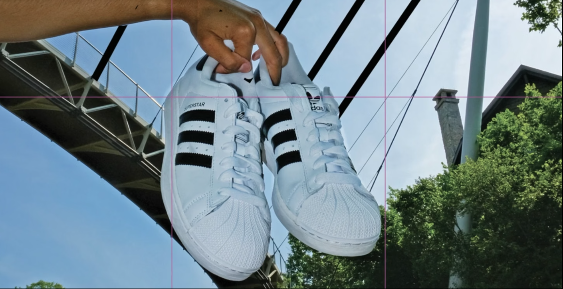 A hand holds a pair of white adidas Superstar sneakers beneath a bridge with trees and blue sky in the background.